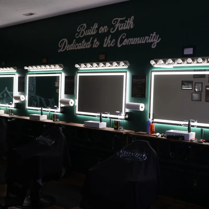 The row of barber chairs at The Greenfield Barbershop lit by vanity bulbs against the deep green wall