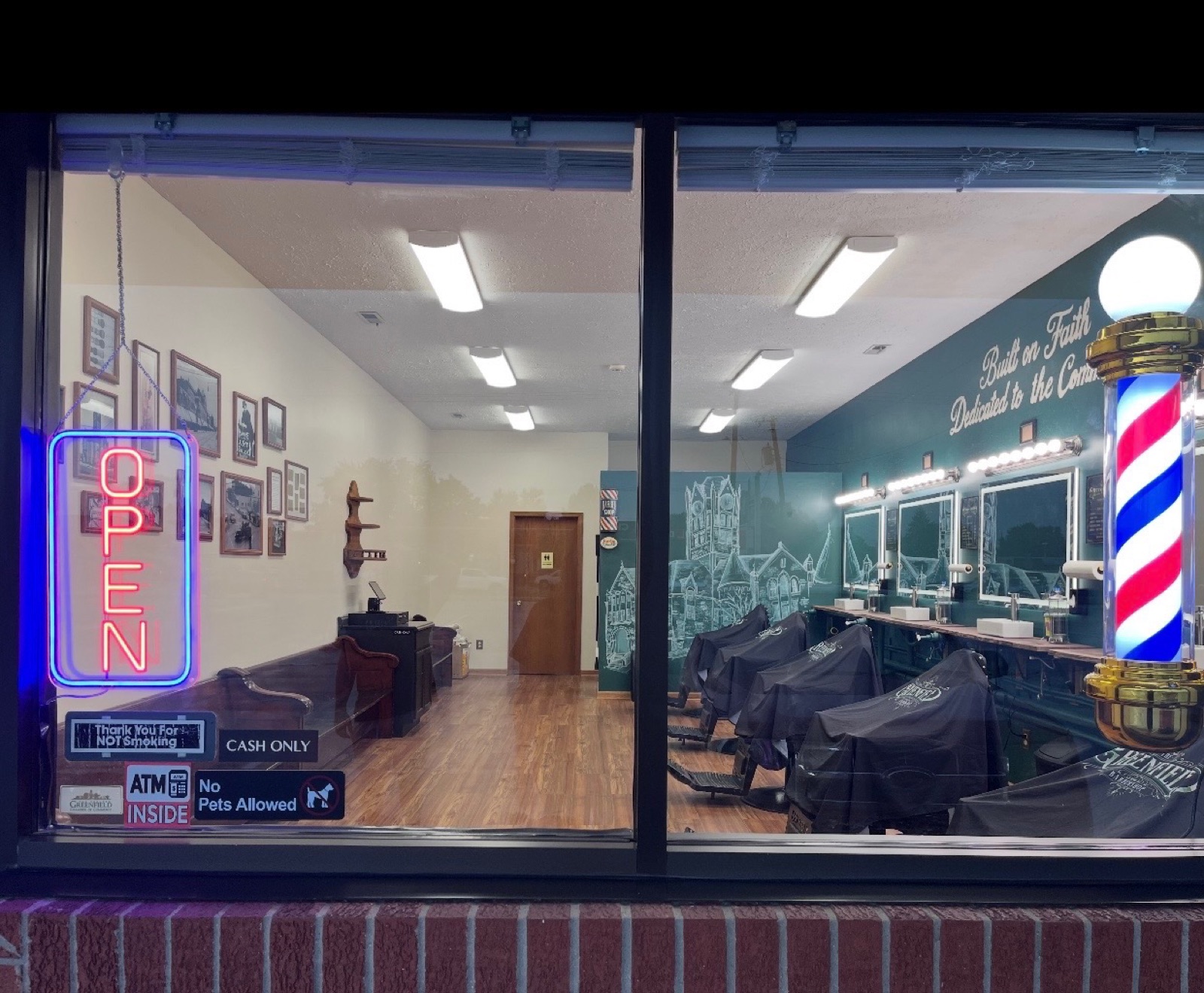 The whole shop seen through the front window at night, neon Open sign lit, barber pole glowing, and the Cash Only and ATM Inside signs in the glass
